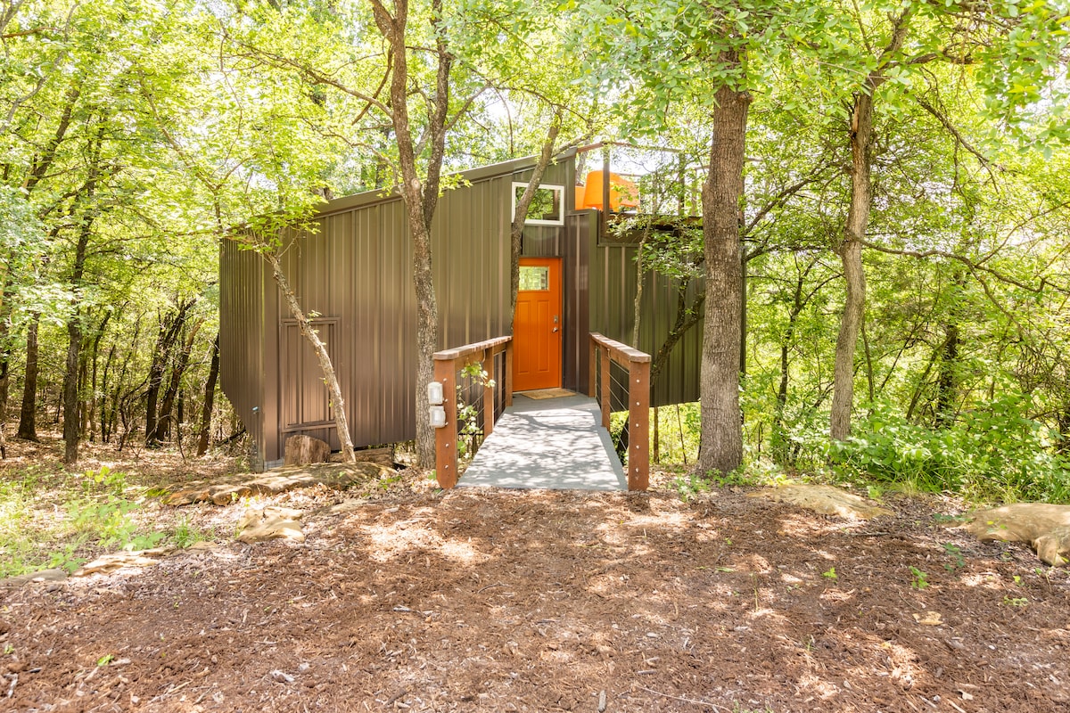 A modern treehouse entrance is framed by lush greenery, featuring a sturdy wooden ramp leading to a vibrant orange front door. The surroundings include a natural forest setting with various trees, enhancing a tranquil ambiance.