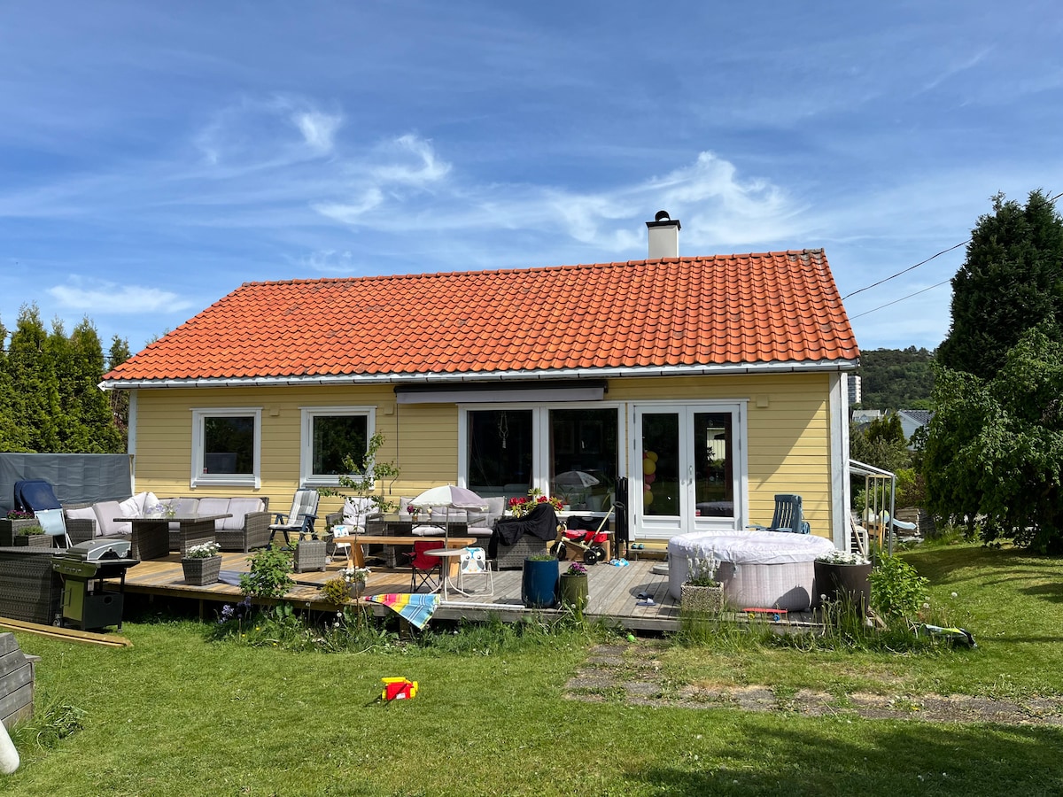 A single-story house is depicted with a yellow exterior and a red-tiled roof. Large glass doors provide views of the deck, which is surrounded by outdoor furniture. A spacious yard is visible, featuring greenery and scattered items, indicating a relaxed, family-friendly environment.