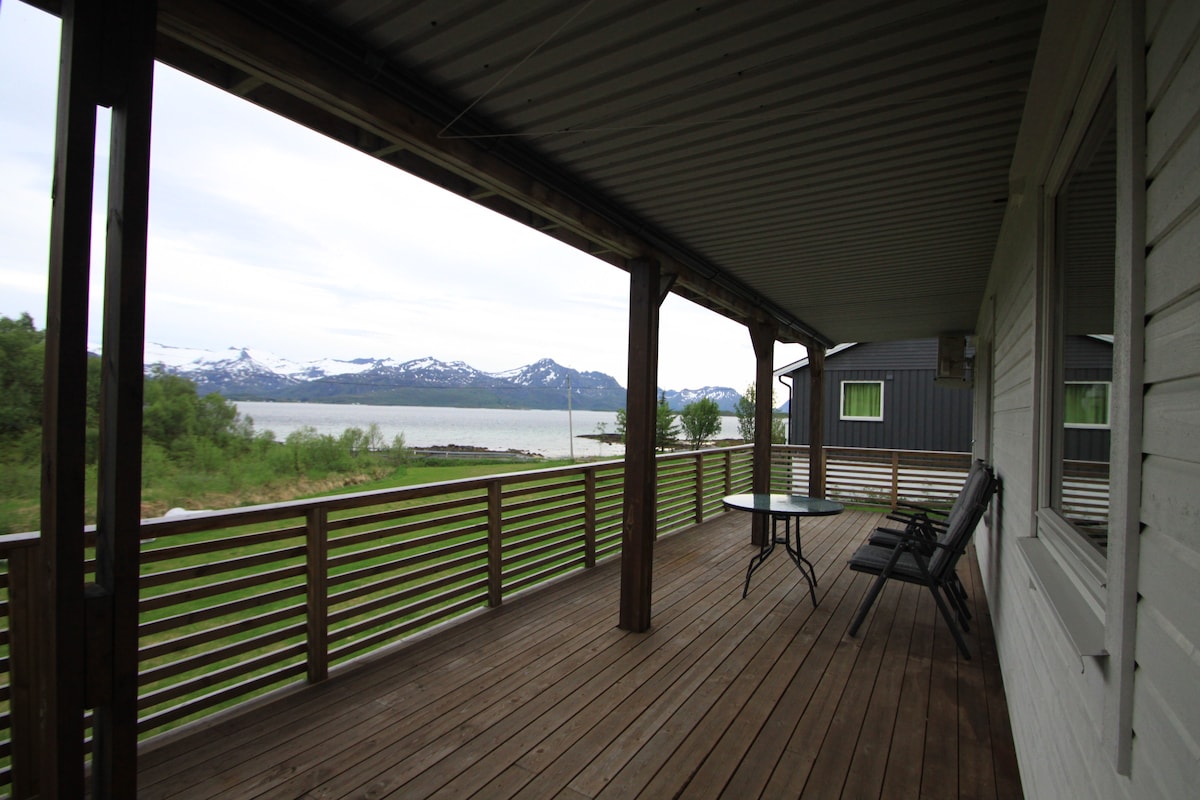 A spacious outdoor deck is visible, featuring a wooden floor and a seating area with two chairs and a small table. The surrounding landscape includes lush greenery and a view of distant snow-capped mountains, with a body of water in the foreground.