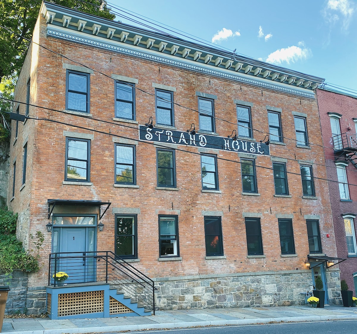 The exterior of the Strand House is depicted, showcasing the historic brick façade with a classic sign. Large black-framed windows are evenly spaced along the front, and a set of steps leads to the main entrance. Flowers add a touch of color near the entrance.