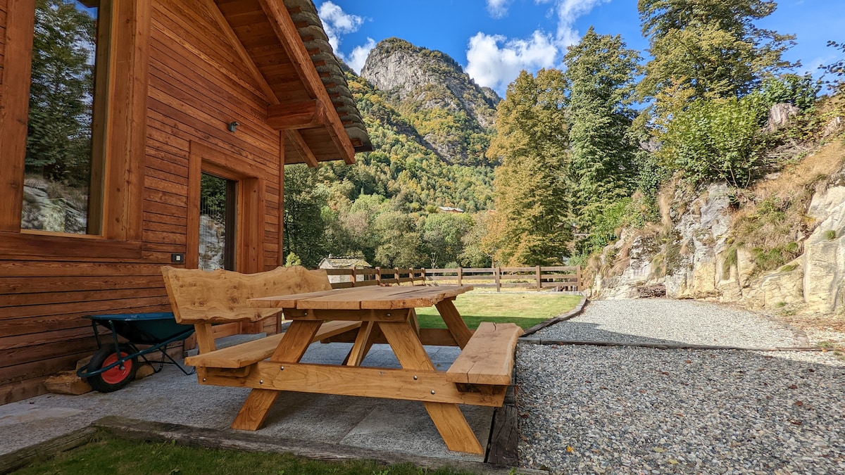 A wooden picnic table is positioned outside the chalet, surrounded by a grassy area and framed by rocky terrain. The backdrop features lush green hills under a partly cloudy sky, emphasizing the serene natural environment.