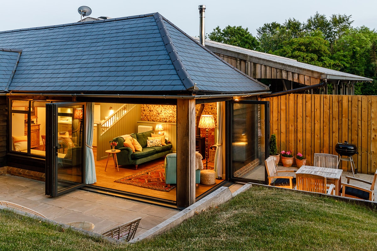 A glimpse of the open-plan living area reveals a comfortable sofa and armchair, with light streaming through large bi-folding doors. Outdoor seating is arranged on the patio, providing a space for al fresco dining and relaxation, framed by well-maintained grass and wooden fencing.