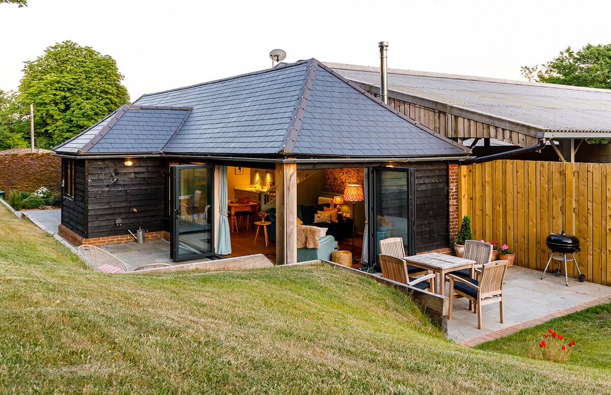 The exterior of The Hovel showcases a wooden cottage with a sloped roof, surrounded by a gentle green slope. Large bi-folding doors invite natural light into the open-plan living space, while a patio area features a dining table and a barbecue for outdoor dining.
