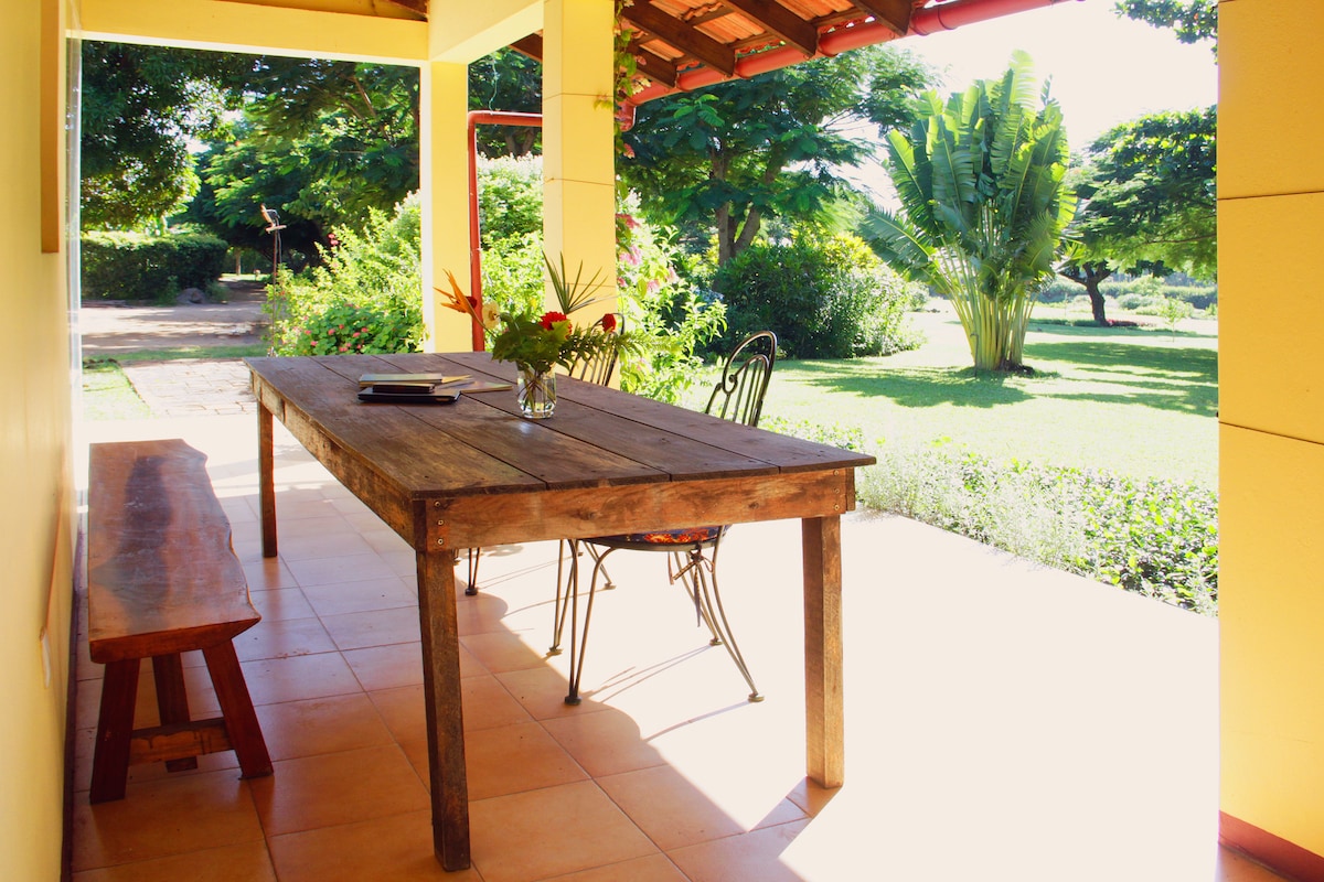 A covered outdoor patio features a rustic wooden table surrounded by four wire chairs. Natural light illuminates the space, highlighting a small vase with flowers on the table. Lush greenery and trees are visible in the background, contributing to a peaceful garden setting.