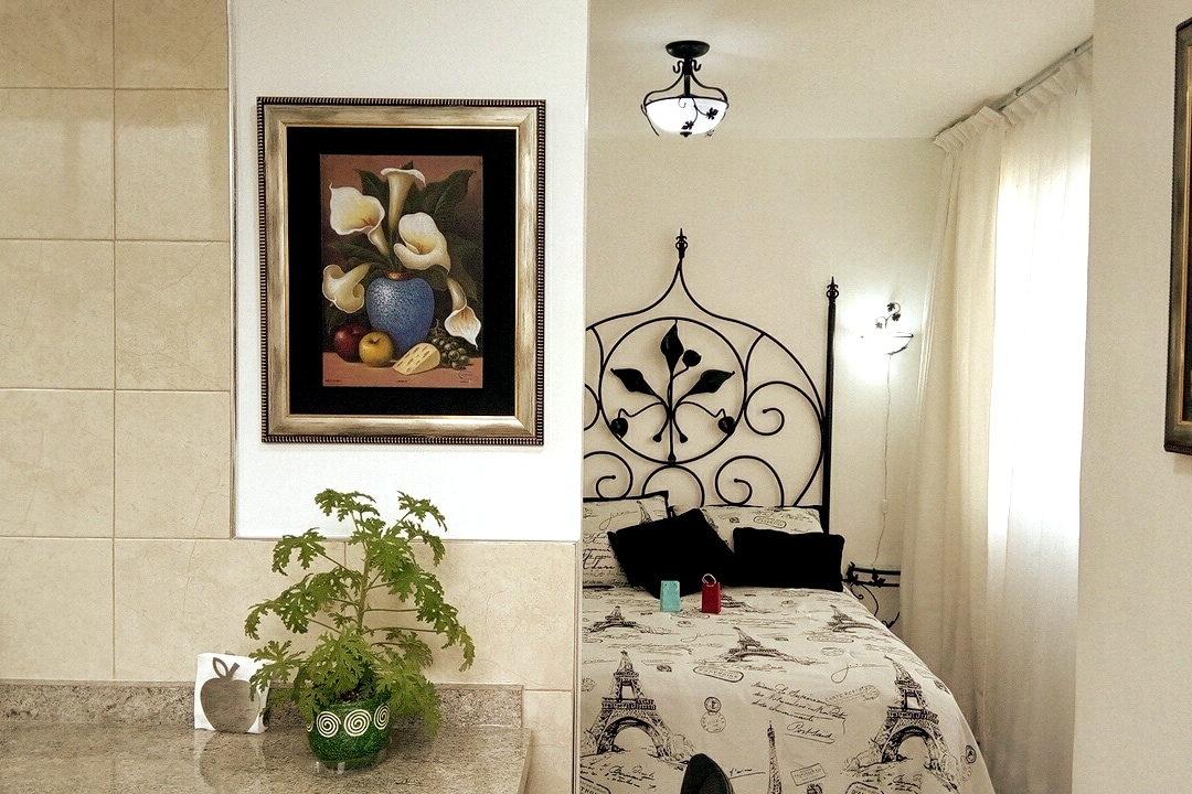 A cozy space features an artistic wall hanging above a decorative bed frame, complemented by light-colored bedding. A small plant adorns the countertop, adding a touch of greenery. Natural light filters through sheer curtains, enhancing the comfortable atmosphere.
