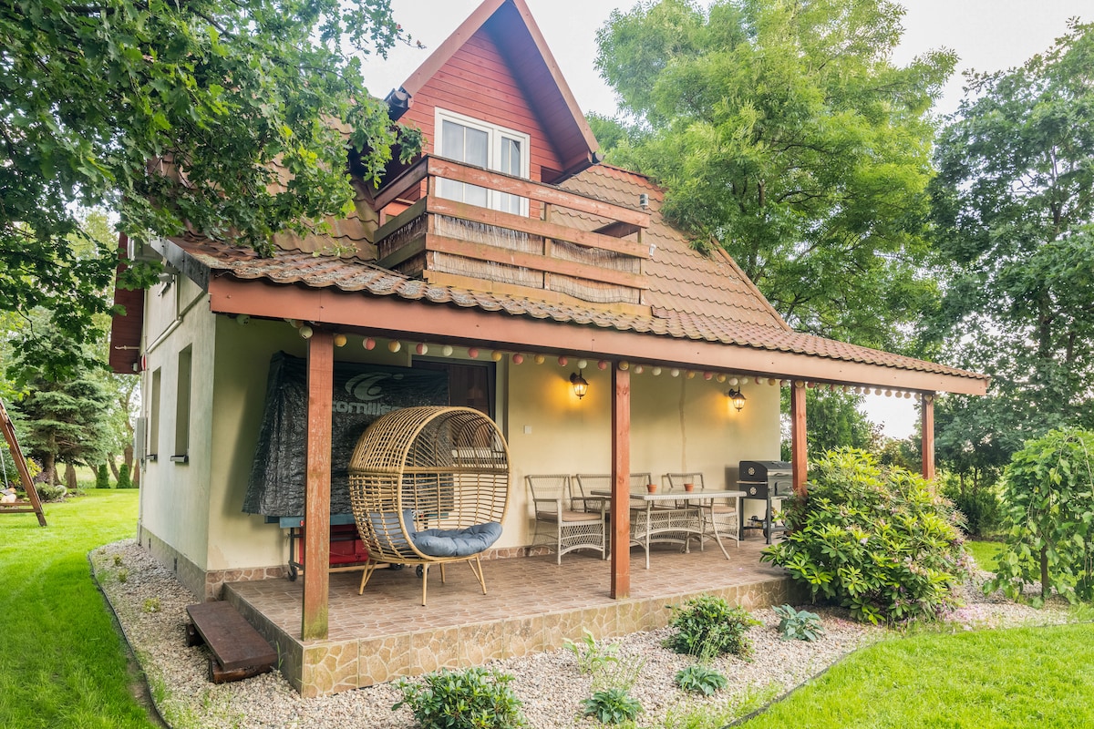 The exterior of the house features a two-story design with a sloped roof and a wooden balcony. A covered patio area includes a hanging chair and several outdoor seating options. Lush green grass and decorative plants enhance the surroundings, creating a peaceful outdoor space.