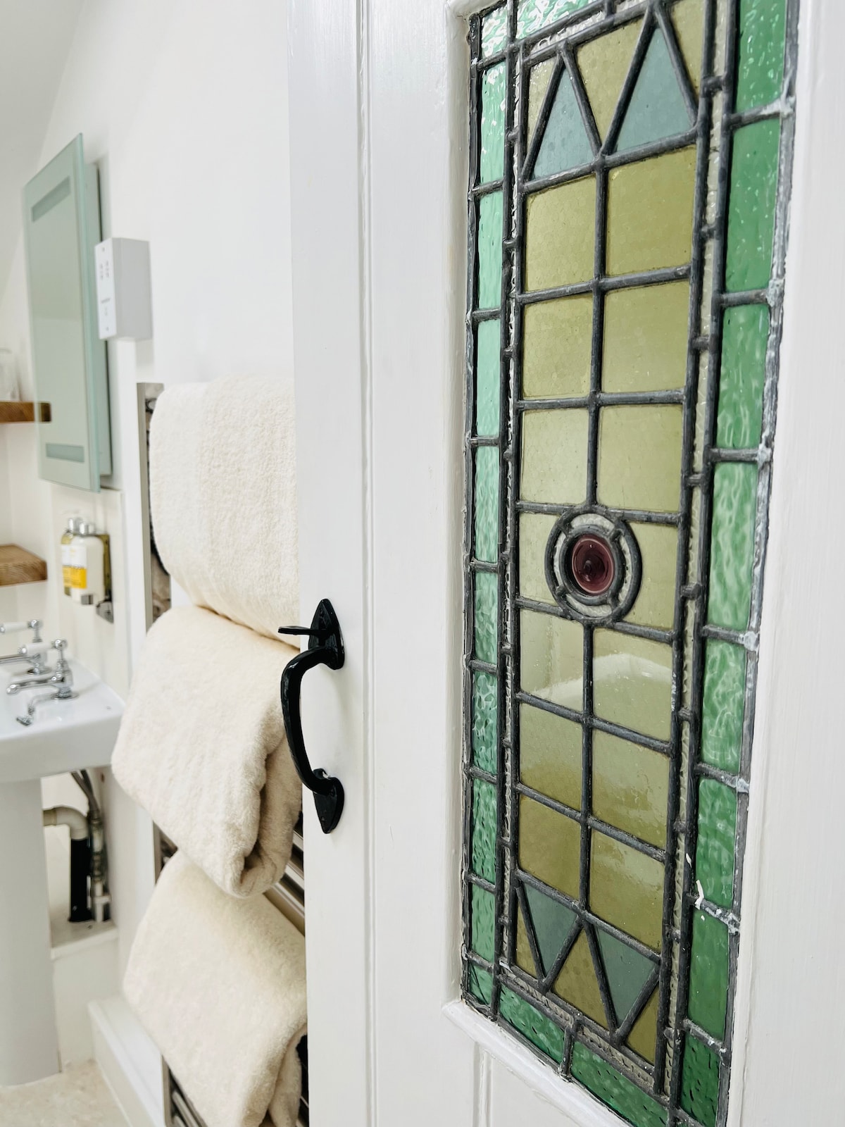 A doorway is featured, adorned with decorative stained glass highlighting green and yellow shades. Fluffy towels are neatly arranged on a rack nearby, while a compact sink is visible, adding functionality to the space.