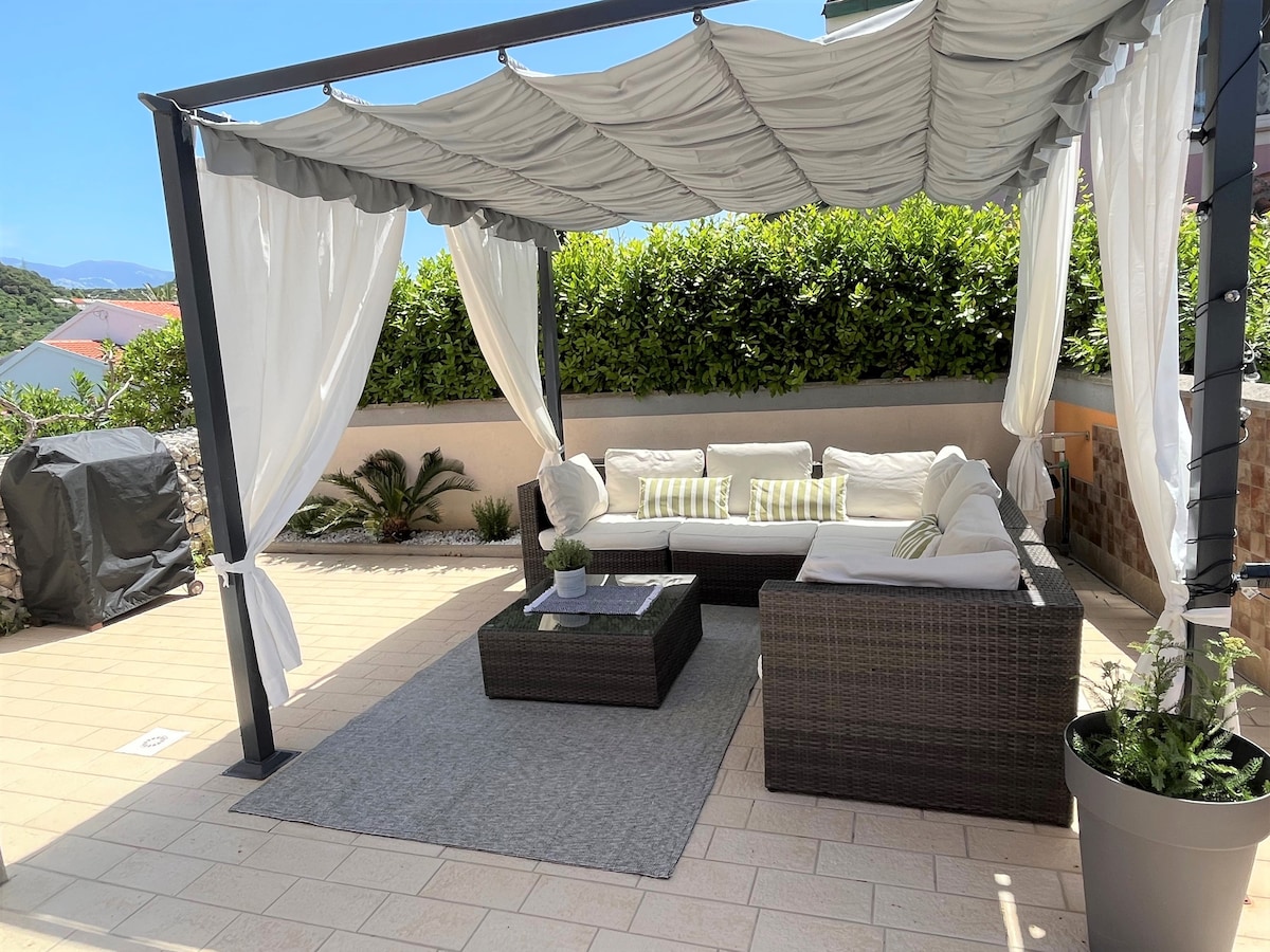 A shaded outdoor lounging area is highlighted, featuring a cozy sectional sofa with light cushions and a low coffee table. Soft drapes frame the space, while greenery surrounds the patio, creating a serene environment for relaxation.