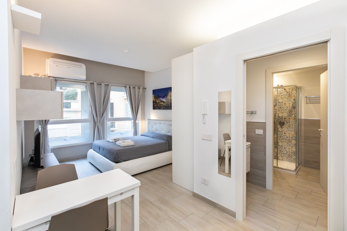 A bright and airy studio apartment is displayed, featuring a spacious bed with a neatly arranged duvet. A small desk is positioned beside the bed, and large windows are dressed with sheer curtains. A modern bathroom with a glass shower is visible through an open door.