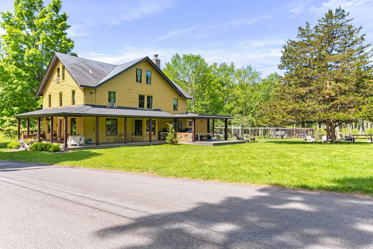 The exterior of a yellow two-story house is surrounded by lush greenery. A wide porch with seating is visible in front, while a spacious lawn leads toward trees and open areas in the background. The road in front provides easy access to the property.
