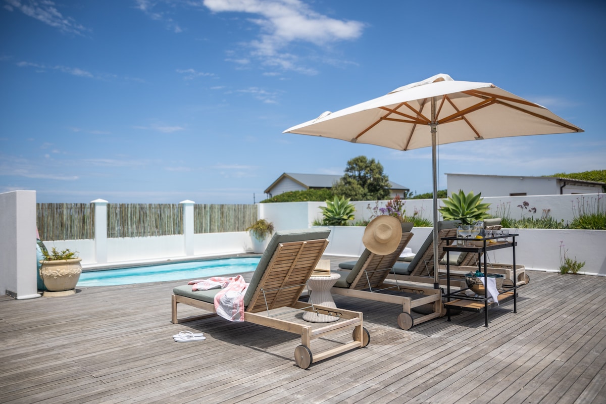 A sunlit deck features two lounge chairs with a large umbrella providing shade. A small side table is positioned next to a comfortable chair, with potted plants lining the edge. The crystal clear pool is visible in the background, framed by a white wall.