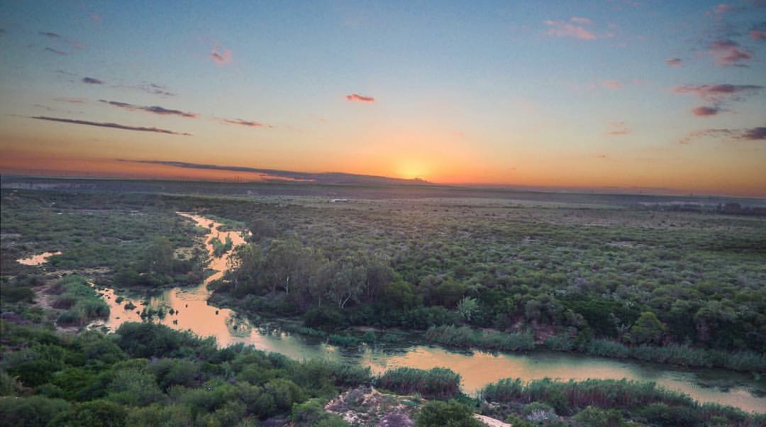 A serene landscape features a winding river surrounded by lush greenery, reflecting the warm hues of a sunset. The sky transitions from soft blues to warm oranges and pinks, creating a gentle backdrop against the expansive horizon.