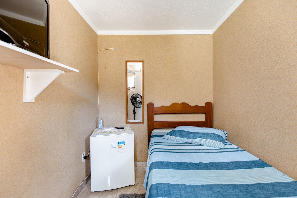 A cozy room features a single bed with a striped blue blanket. A small refrigerator is positioned beside the bed, while a mirror and a television are mounted on the wall. Natural light spills in through a nearby window.