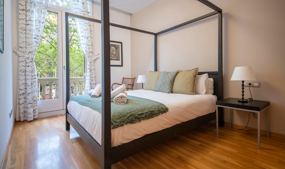 A spacious bedroom features a queen-sized canopy bed adorned with soft pillows and a cozy throw. Light pours in through large windows dressed with sheer curtains, offering views of the tree-lined street. A bedside lamp complements a small table, enhancing the room's functionality.