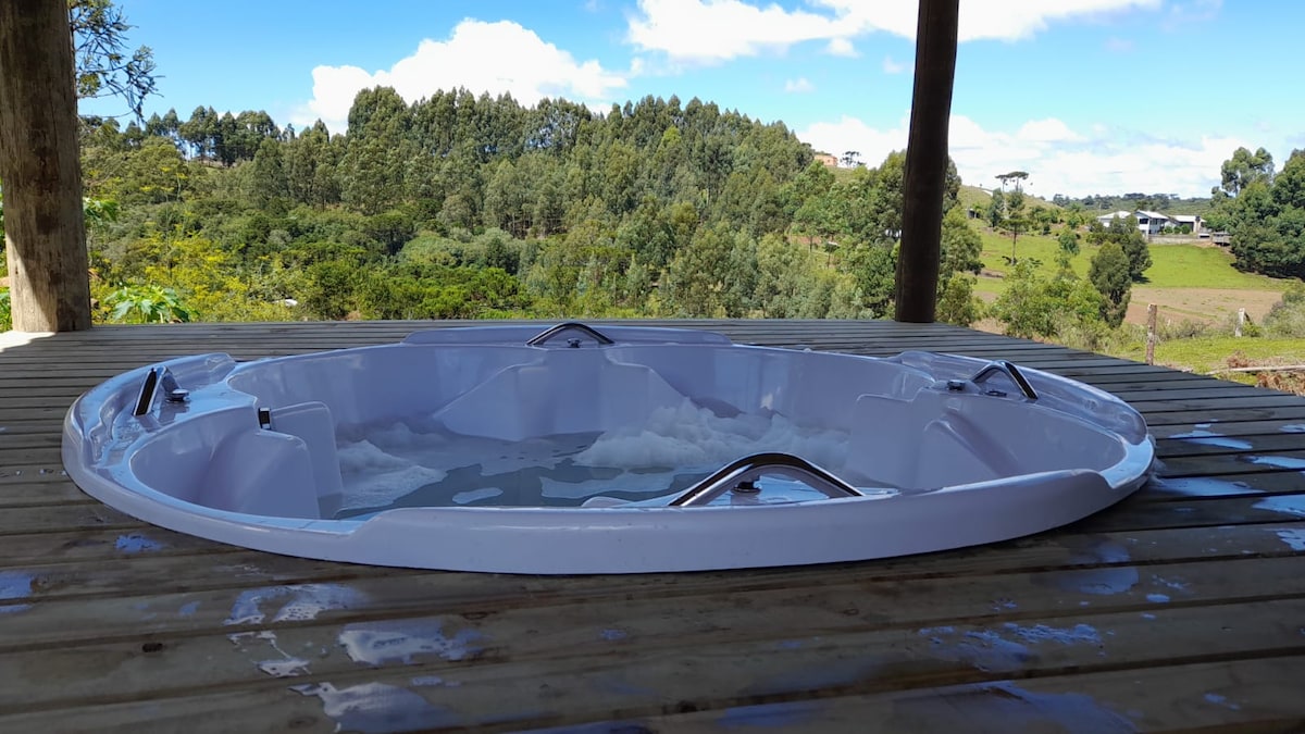 A circular hot tub is positioned on a wooden deck, surrounded by lush greenery and distant hills. The clear sky and soft clouds are visible in the background, creating a serene outdoor setting.