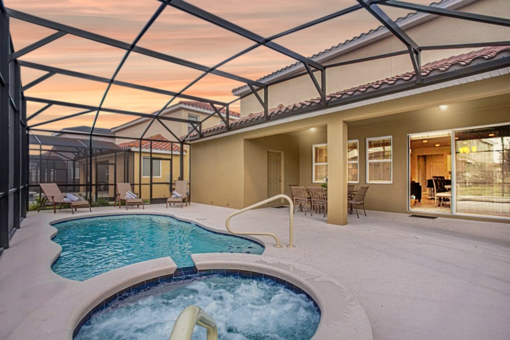 A private pool area features a curvy pool alongside a spa, both protected by a screened enclosure. Sun loungers are arranged around the pool, and the home is visible in the background, showcasing large glass sliding doors that invite outdoor views.