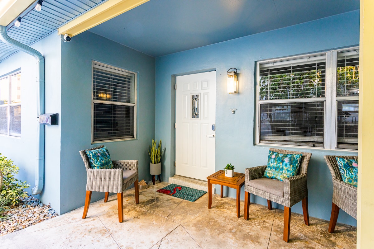 An inviting entrance features a light blue exterior wall. Two wicker chairs with colorful cushions are positioned beside a small wooden table. A welcome mat is placed in front of the door, which is framed by wall sconces and surrounded by lush greenery.