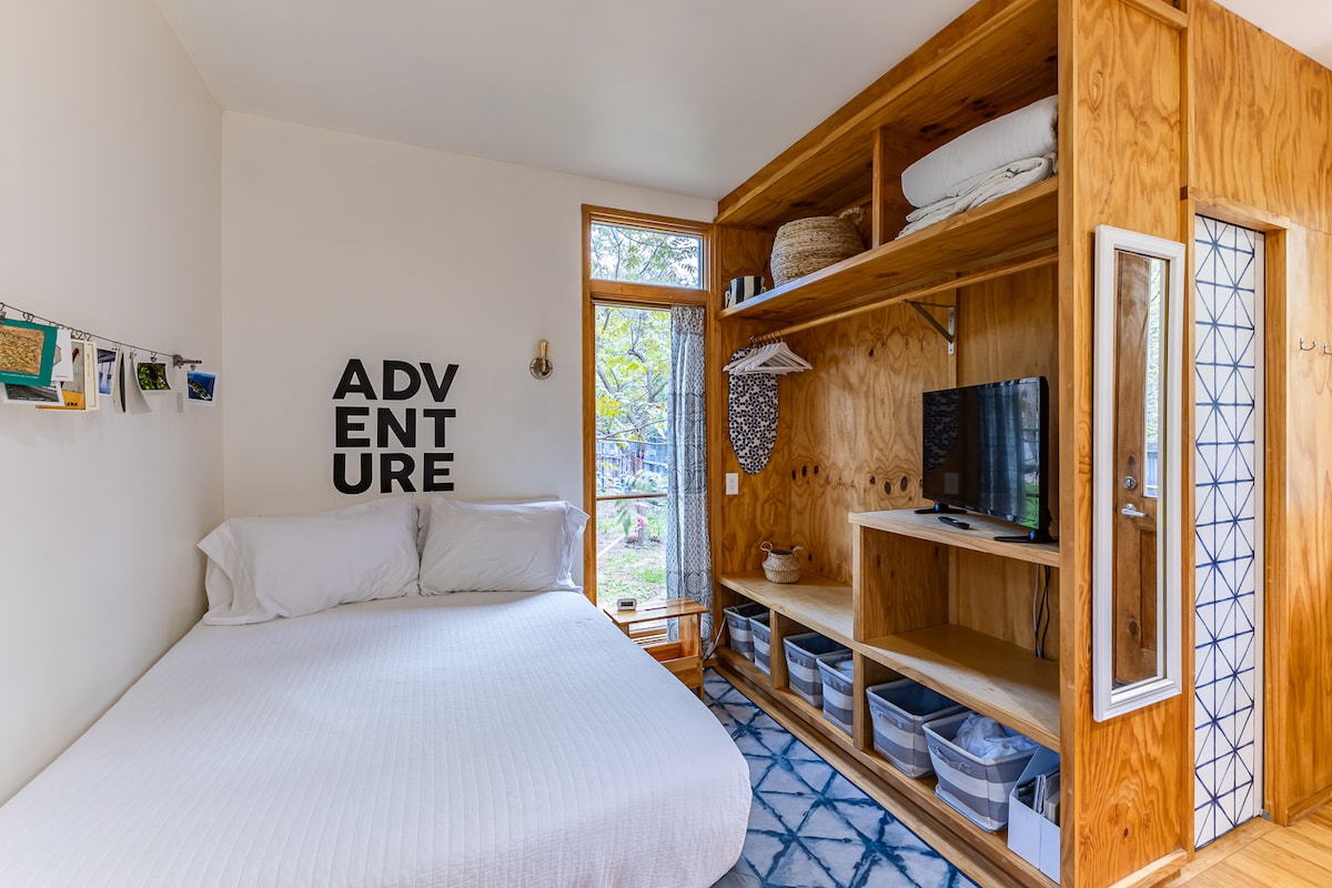 A bright sleeping area features a double/full bed dressed in neutral linens, with an accent wall reading 'ADVENTURE.' Natural light enters through a nearby window, illuminating wooden shelving that holds neatly organized baskets and a small television.