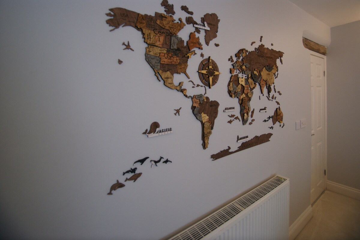 A decorative wooden world map is displayed on a light grey wall, featuring various countries highlighted in different shades. The map includes a compass rose and features small animal silhouettes, with a radiator visible beneath it. A white door is seen at the end of the hallway.