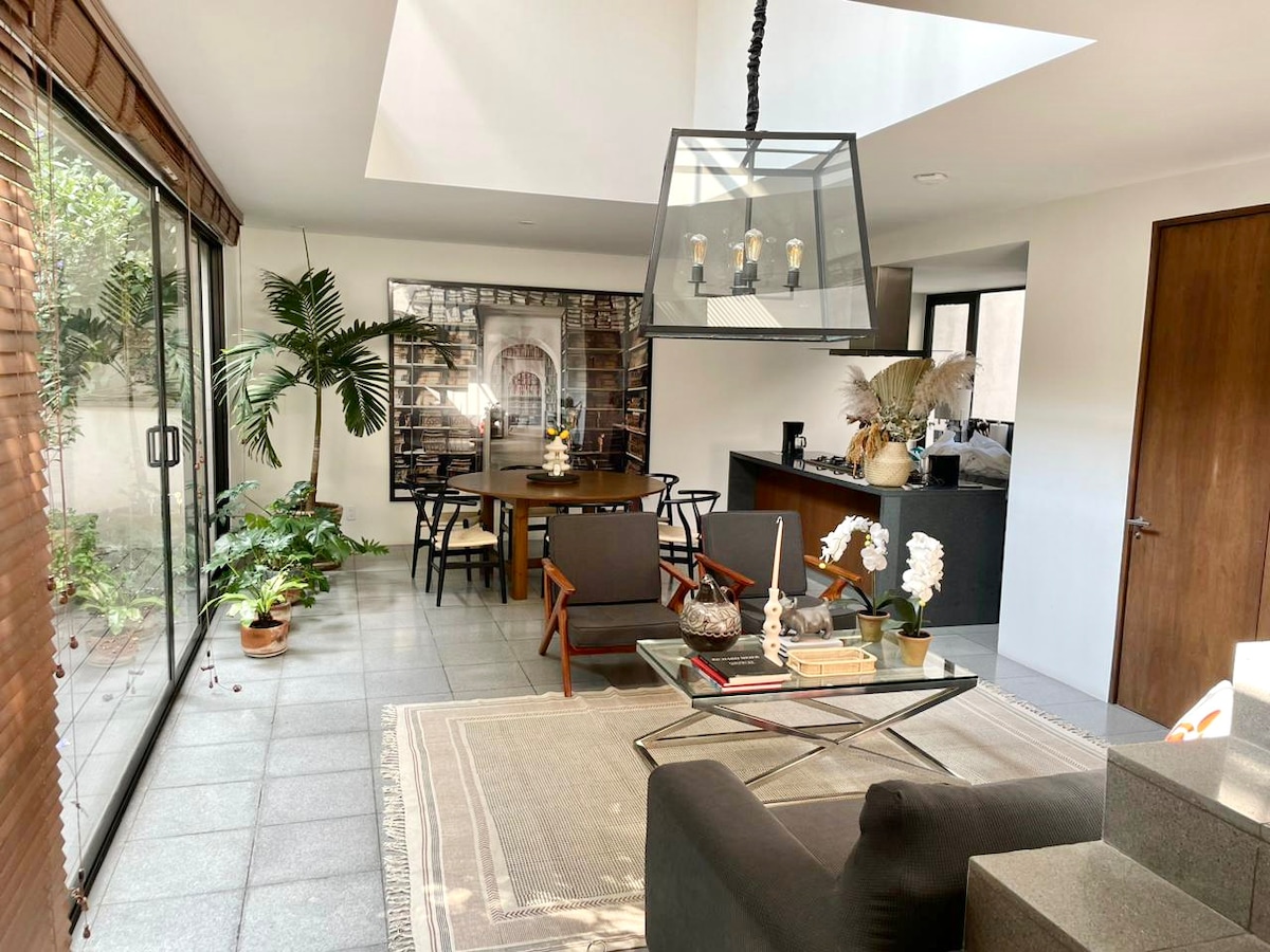 An open living and dining area showcases a round dining table surrounded by black chairs and potted plants. Large glass doors lead to the leafy patio, while a modern pendant light hangs above the dining space. A cozy seating arrangement includes a sleek coffee table and decorative accents.