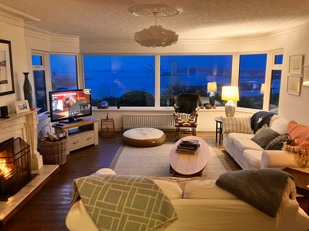 A cozy living room features a large bay window offering views of Whiterock Bay. Soft seating arrangements are complemented by a warm fireplace. A television is positioned in the corner, and a circular ottoman sits in the center. Light fixtures create a relaxed atmosphere.