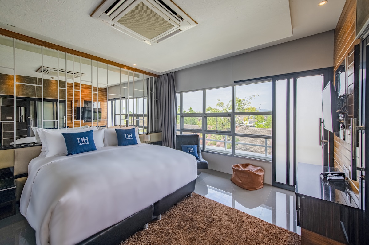 A bright and spacious bedroom features a large bed adorned with white linens. Floor-to-ceiling windows allow natural light to fill the room, offering views of the outdoors. A comfortable seating area includes a lounge chair and a small table. Modern decor highlights the inviting atmosphere.