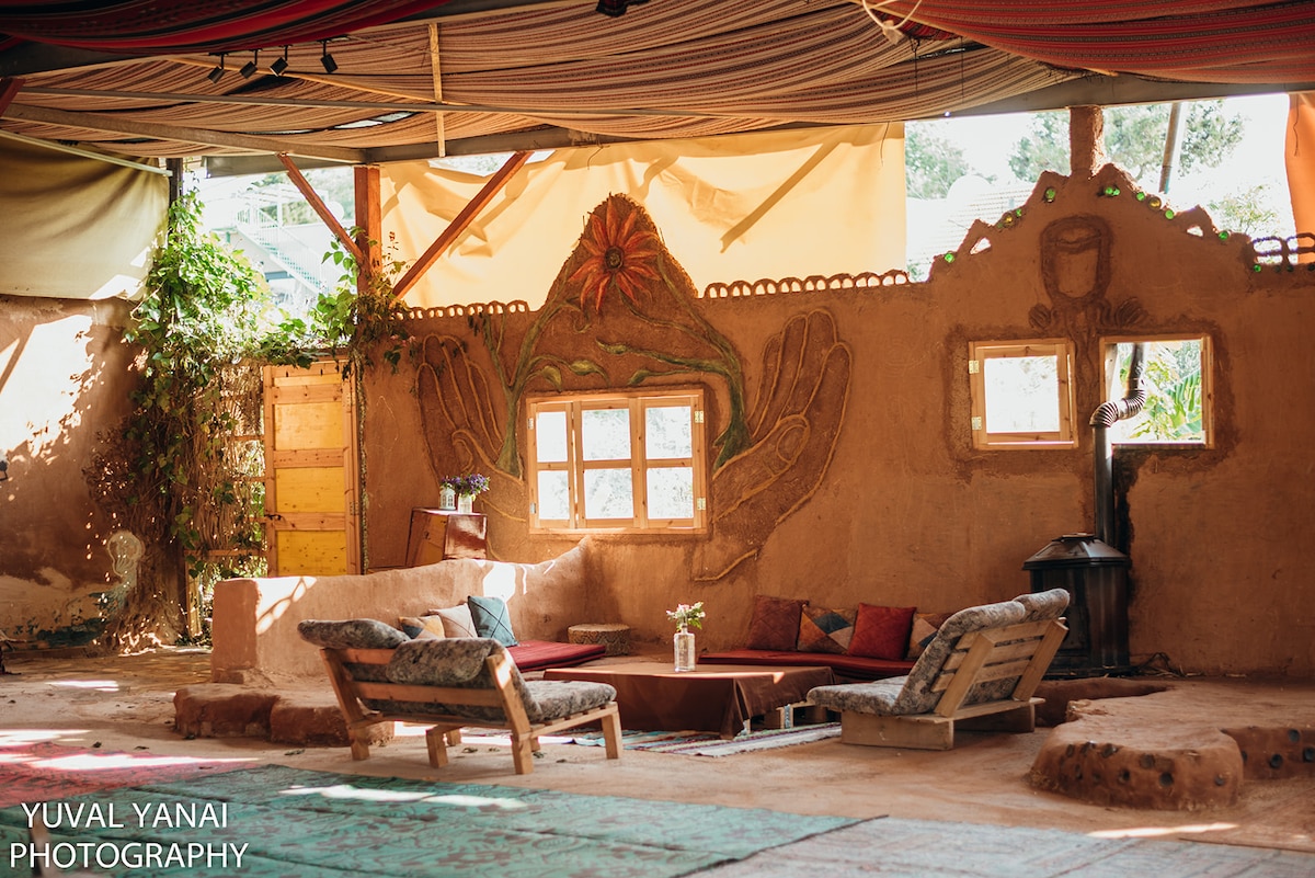 An open communal area is highlighted by earth-toned clay walls featuring artistic hand motifs. Natural light filters through skylights and window openings, illuminating wooden seating arrangements with colorful cushions. Greenery decorates the space, enhancing the rustic vibe of this handmade mud-building.