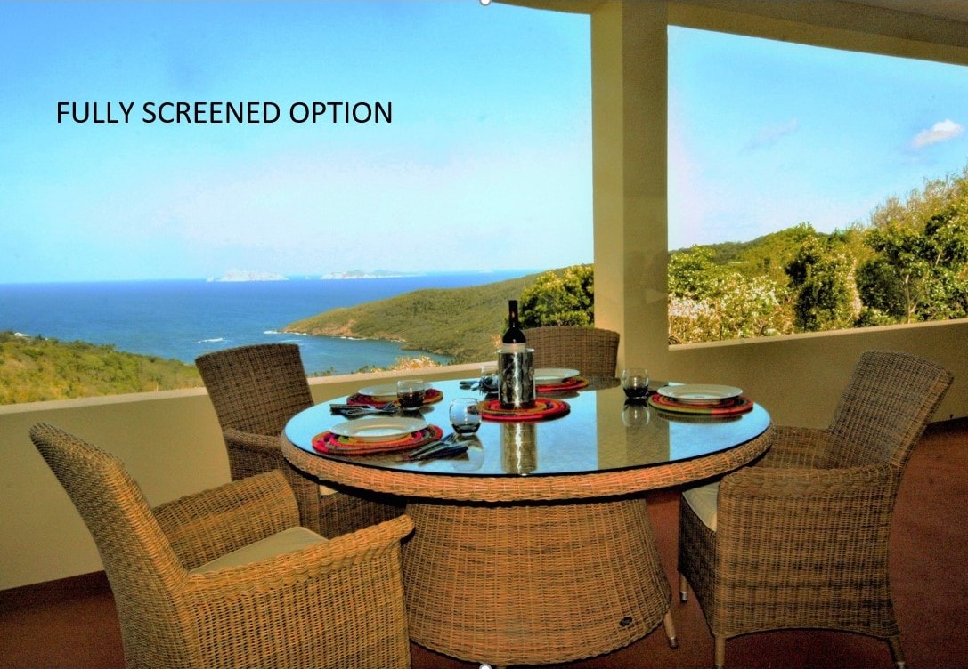 A screened outdoor dining area features a round table surrounded by four wicker chairs, set against a backdrop of sweeping coastal views. The table is arranged with colorful plates and a wine bottle, offering a serene setting for meals.