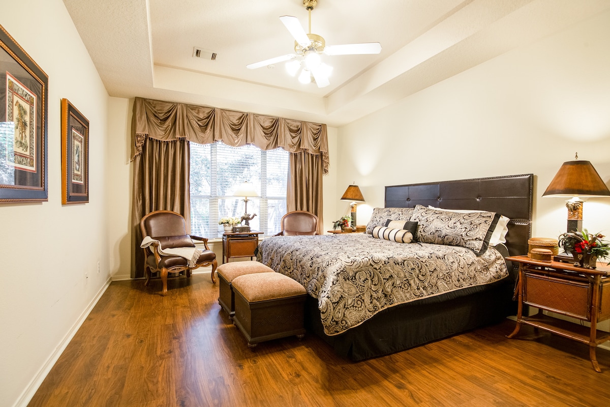 A spacious bedroom features a large bed with decorative bedding and multiple accent pillows. Two side tables with lamps are positioned beside the bed, and a cozy bench sits at the foot. Natural light enters through a large window, partially covered by elegant curtains.
