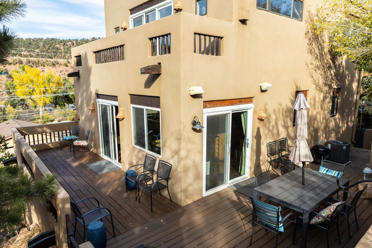 An expansive deck is visible, featuring a large outdoor dining table with several chairs and an umbrella. Sliding glass doors lead inside, while the surrounding area showcases trees and colorful autumn foliage, enhancing the natural setting.