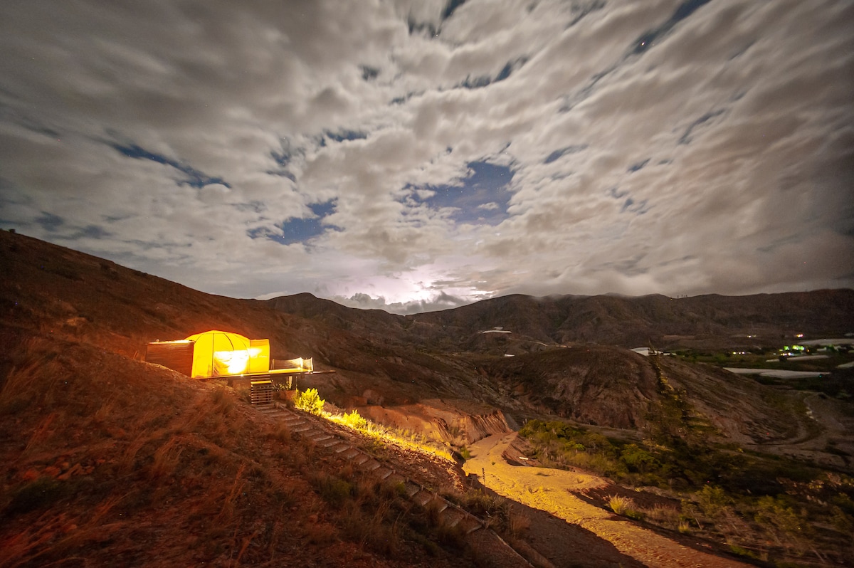 An orange glamping tent is positioned on elevated ground, surrounded by a rugged mountainous landscape. Soft lighting from the tent creates a warm glow against the night sky, which is filled with textured clouds. The winding path leads into the distance, inviting exploration of the serene environment.