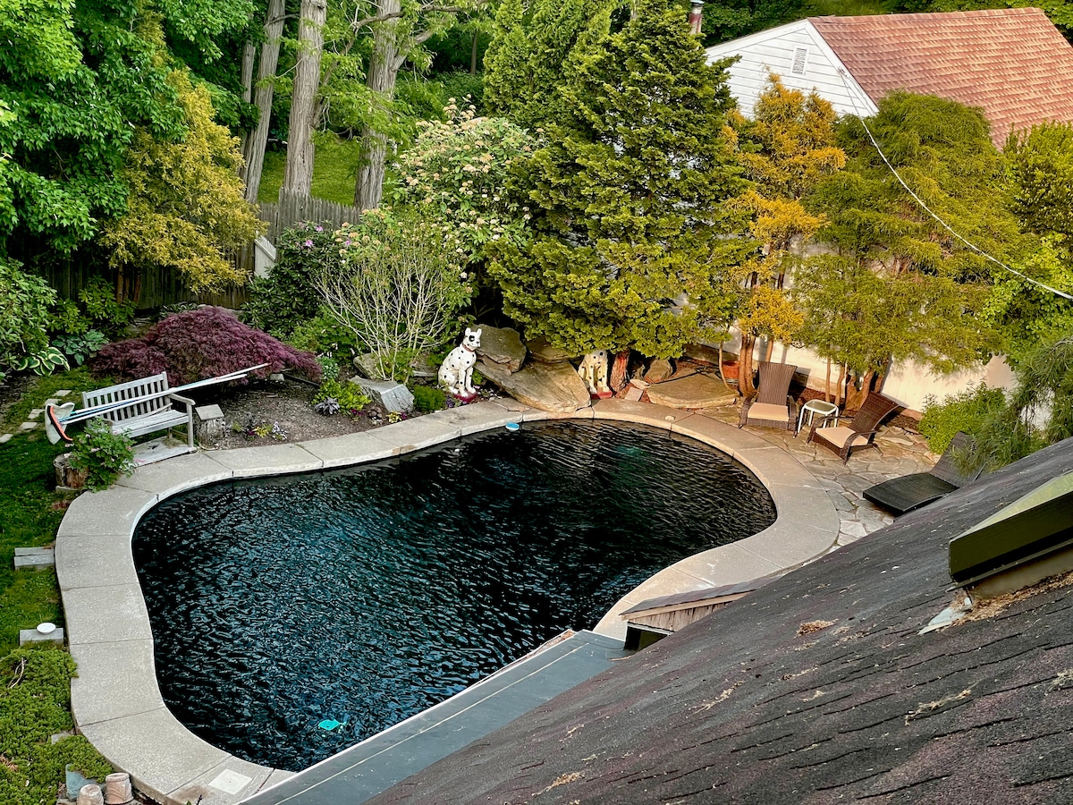 A spacious outdoor area features a curvilinear pool surrounded by natural greenery and landscaped stonework. Sun loungers and a shaded seating area are positioned nearby, providing relaxation options. The pool's dark surface reflects the surrounding foliage, creating a serene retreat atmosphere.