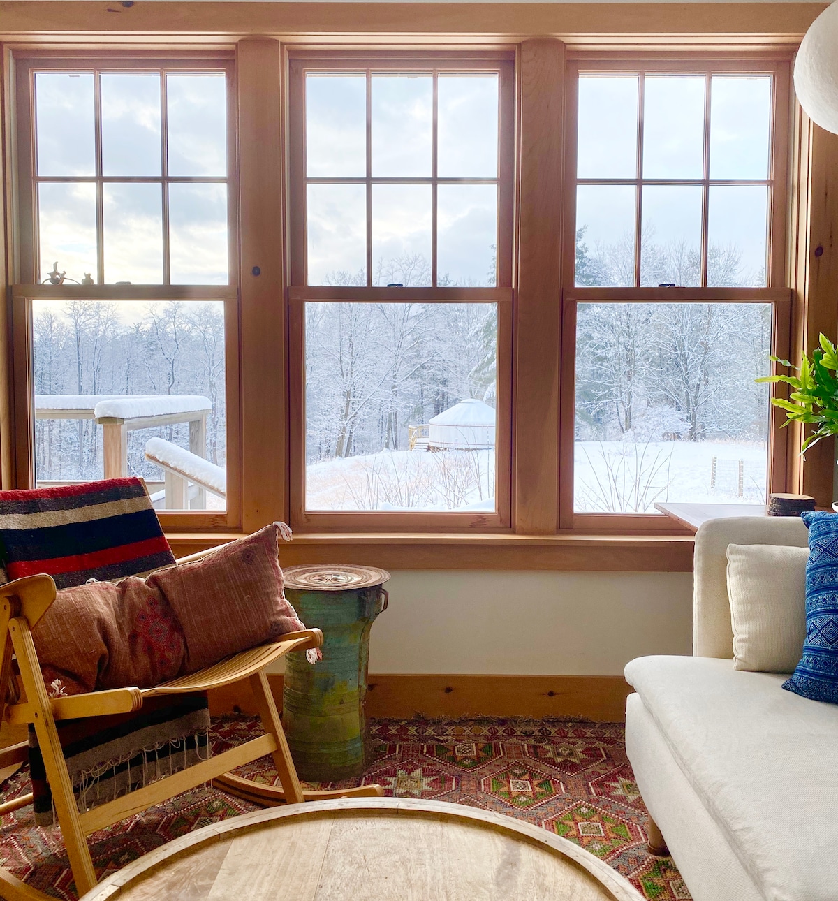 A cozy seating area features a white couch and a wooden chair adorned with colorful cushions. Large windows showcase a winter landscape, with snow-covered ground and trees visible outside. Light enters the room, highlighting the warm tones of the wooden frames and the inviting rug.
