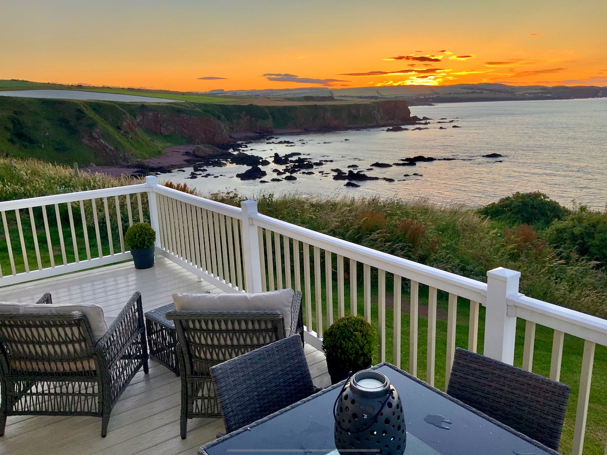 A deck area features outdoor furniture overlooking a tranquil sea, with a stunning sunset casting warm hues over the coastline. Lush greenery frames the scene, enhancing the serene atmosphere of the location.