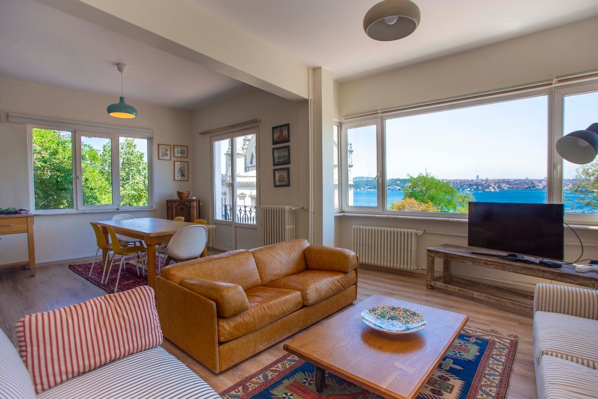 A modern living area is depicted, featuring large windows that frame views of the Bosporus. Comfortable leather sofas are arranged around a central coffee table. A dining table sits nearby, accompanied by white chairs, while a television is positioned in the corner, enhancing the cozy atmosphere.