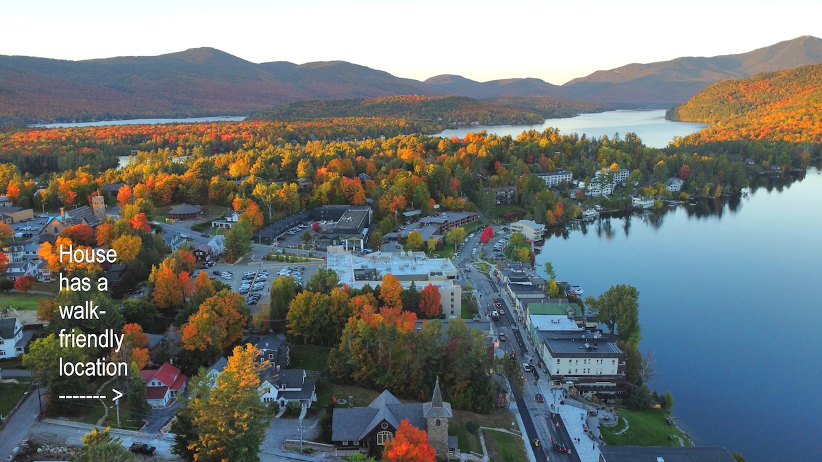 An aerial view highlights a vibrant autumn landscape surrounding the home, with colorful leaves covering trees and the nearby hills. The town below shows shops and amenities within walking distance, while a tranquil lake reflects the surrounding mountains and foliage.
