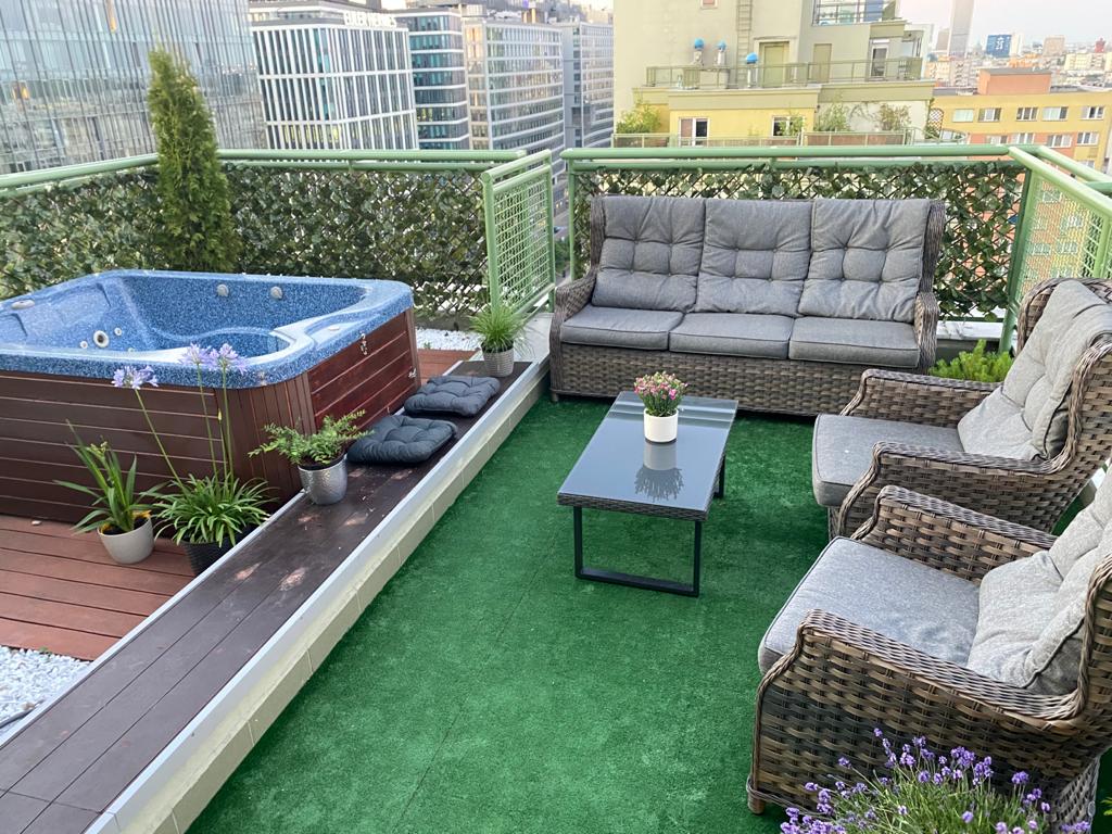 A rooftop terrace features a hot tub surrounded by greenery. Comfortable seating is arranged around a glass-top table, and potted plants add natural accents. Soft green grass covers the floor, blending with the urban backdrop of city buildings.