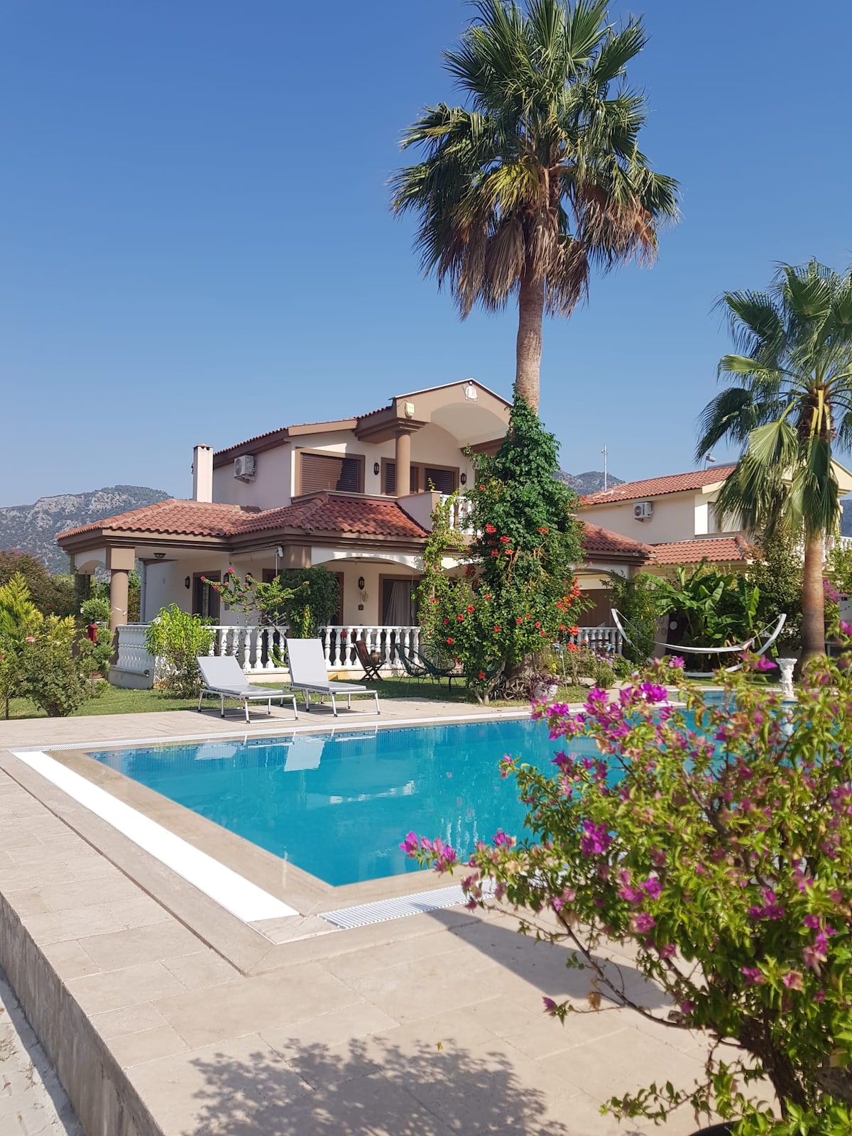 A serene villa with a tiled roof is surrounded by lush greenery and vibrant flowers. A private pool reflects the clear blue sky, while comfortable lounge chairs are positioned on the pool deck, inviting relaxation in a peaceful outdoor setting.