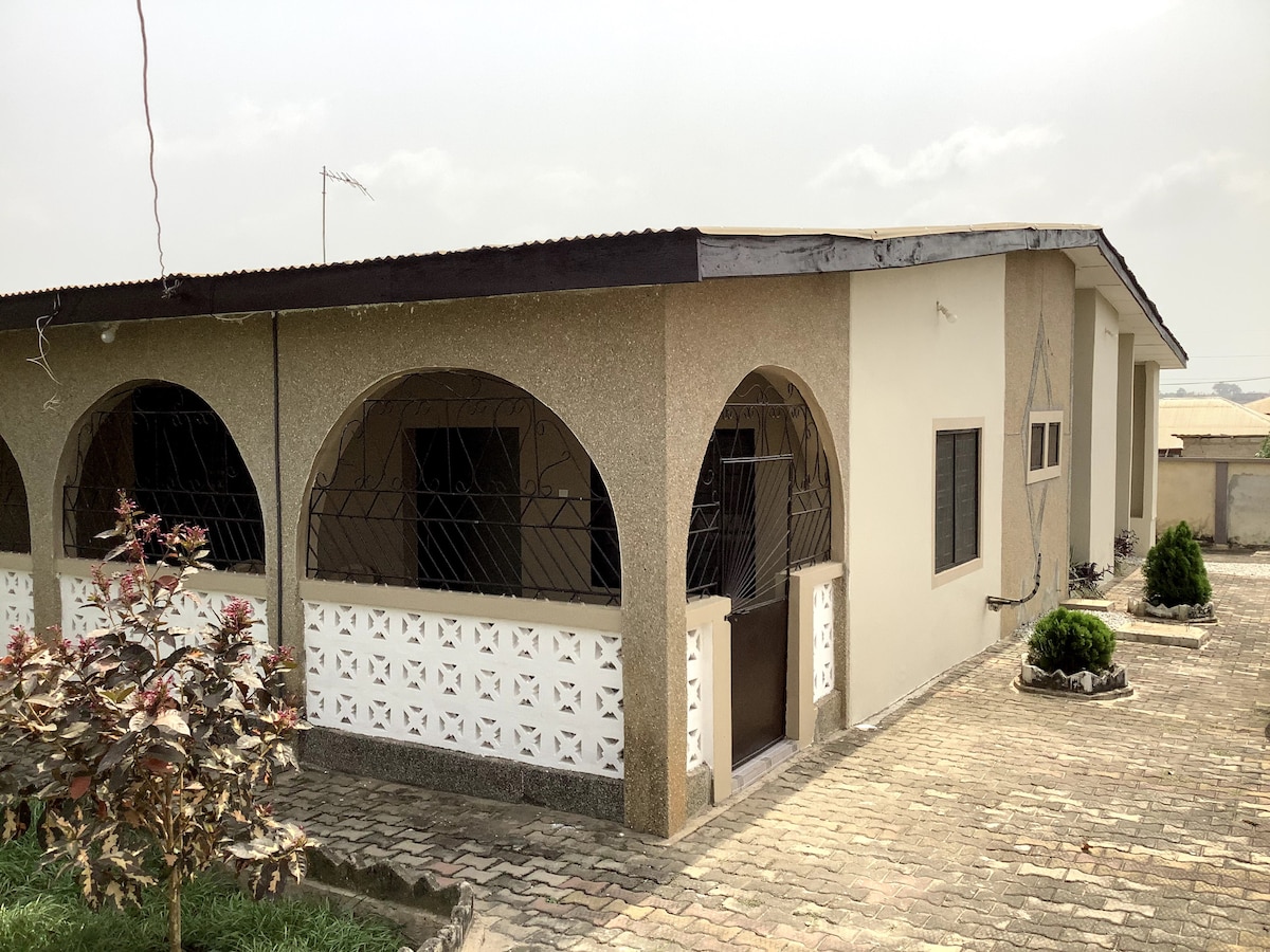 The exterior of the house features a modern design with an arched entrance and decorative brickwork. The paved drive leads to a well-maintained garden area, framed by shrubs and small plants, under a gently cloudy sky.
