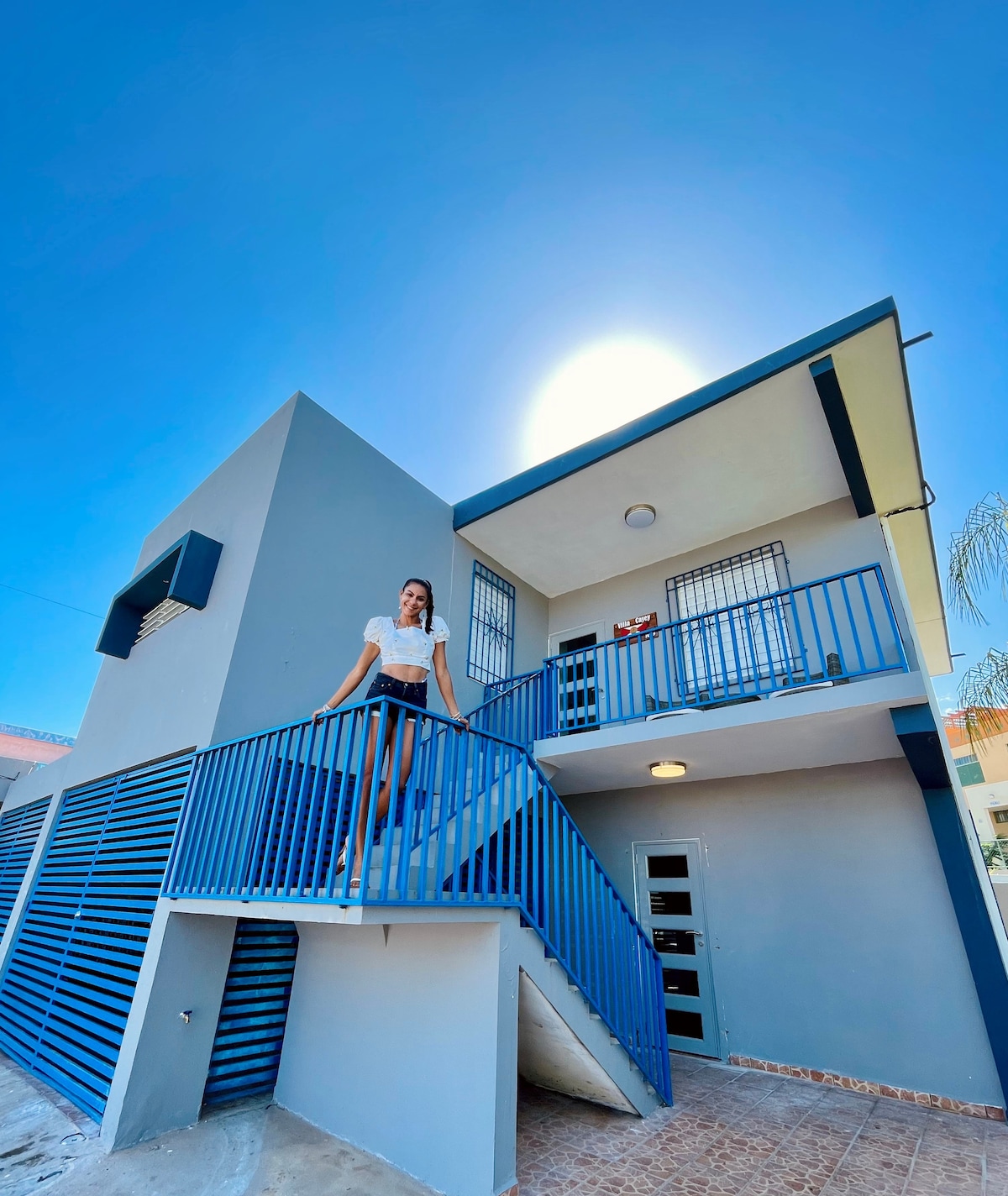 A two-story house is depicted, featuring a blue exterior and a spacious staircase with a blue railing leading to the upper level. Sunlight illuminates the scene, creating a bright atmosphere. Window screens can be seen, complementing the modern design.