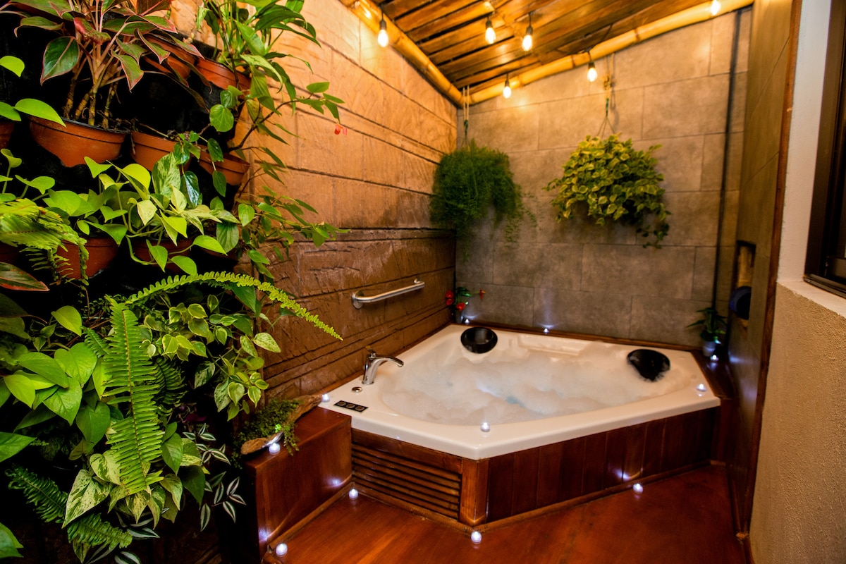 A private jacuzzi is surrounded by greenery and is softly lit by string lights, creating a serene atmosphere. The wooden deck complements the natural elements, while potted plants enhance the tranquil setting. The space invites relaxation after a day of exploration.