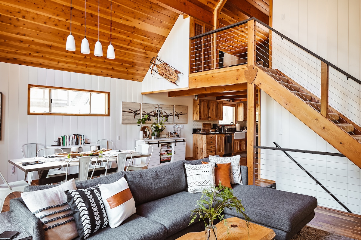 An open living area features a modern sectional sofa with decorative throw pillows, complemented by wooden beams overhead. A dining space with white chairs is visible, along with a rustic kitchen in the background. A staircase leads to the upper level, enhancing the cozy cabin atmosphere.