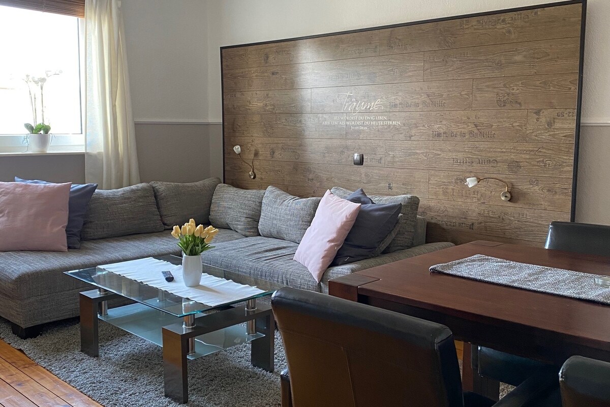 A modern living area features a large sectional sofa adorned with light pink pillows. A glass coffee table reflects light, positioned on a soft area rug. A dining table with black chairs is visible, along with a decorative vase of tulips on the table.