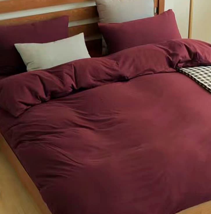 A spacious bed is dressed in warm burgundy bedding, accompanied by several decorative pillows in varying shades. A textured throw blanket adds a layered look. Soft natural light subtly illuminates the room, enhancing the cozy ambiance.