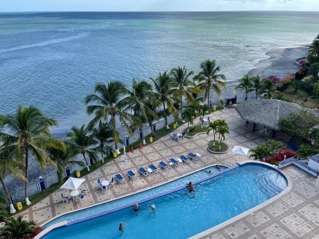 A tranquil swimming pool is seen adjacent to the Pacific Ocean, surrounded by palm trees. Sun loungers are placed along the poolside area, with a thatched-roof gazebo visible in the background. The calm water meets the shoreline lined with greenery, enhancing the serene atmosphere.