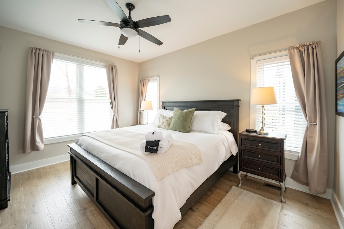 A well-lit bedroom features a spacious bed with crisp white linens and decorative pillows. Soft curtains frame two windows, allowing natural light to fill the room. A nightstand with a lamp and a dresser enhance the cozy atmosphere of this inviting space.