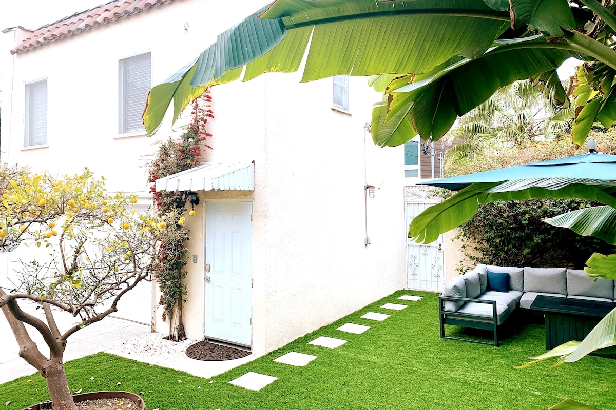 A well-maintained backyard is featured, with a manicured green lawn surrounded by tropical plants. A seating area is positioned to one side, offering a comfortable space for relaxation. A small pathway leads from the door to the lawn, enhancing accessibility.