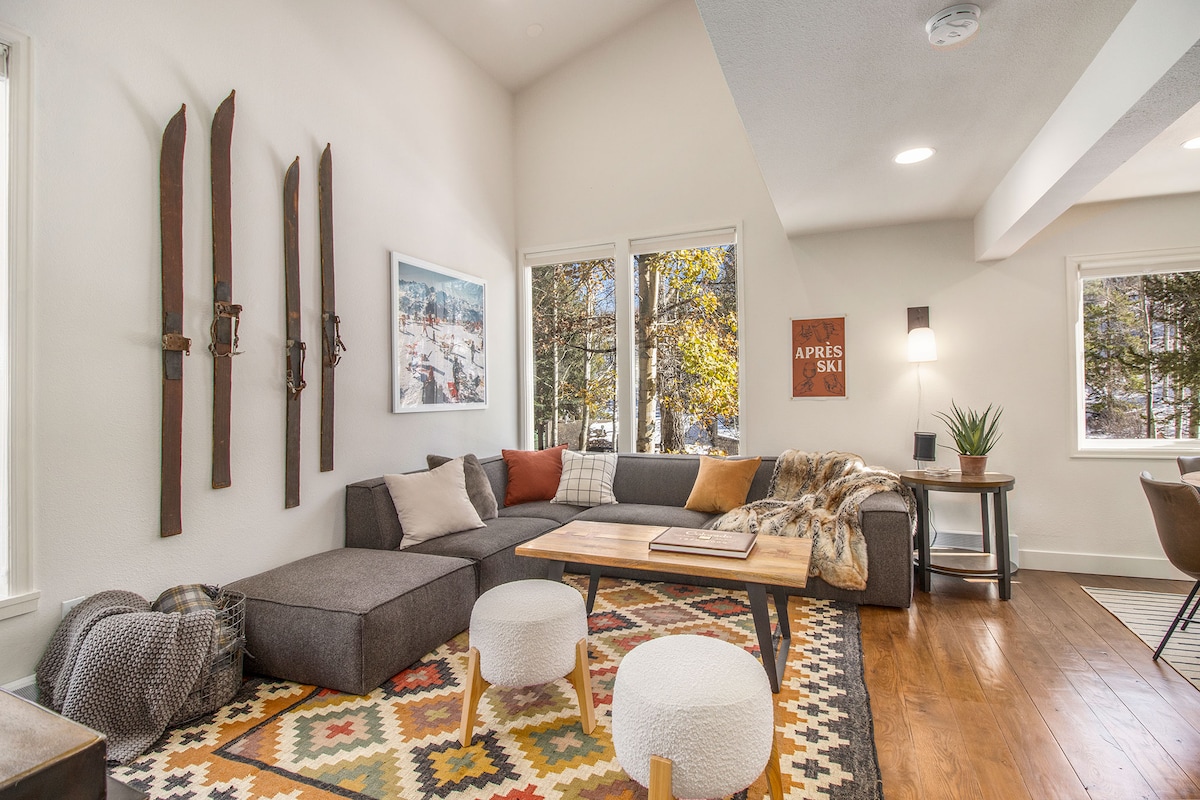 A cozy living space features a large sectional sofa adorned with a soft blanket, complemented by a rustic wooden coffee table. Decorative ski equipment hangs on the wall, and natural light floods in through large windows, illuminating the vibrant area rug beneath.