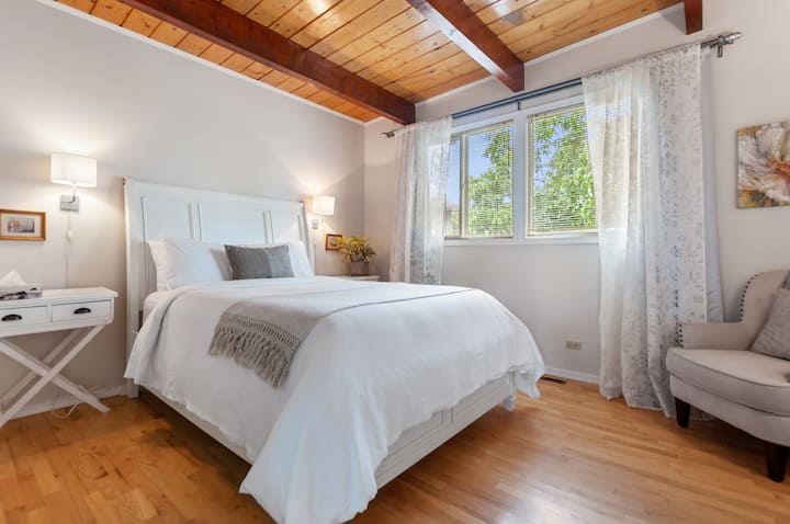 El hermoso dormitorio principal cuenta con vistas al jardín, techo abovedado con vigas de madera, colchón y sábanas gruesas y cómodas, pisos de madera, armario, silla, espejo, cortinas oscuras, cómoda, armario.