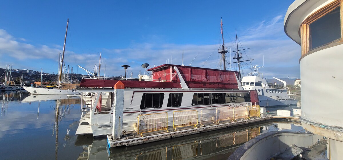 A spacious yacht is docked at a marina, with a striking red roof contrasting against the blue sky. Reflections from the calm water enhance the serene atmosphere, while several other boats are visible in the background.